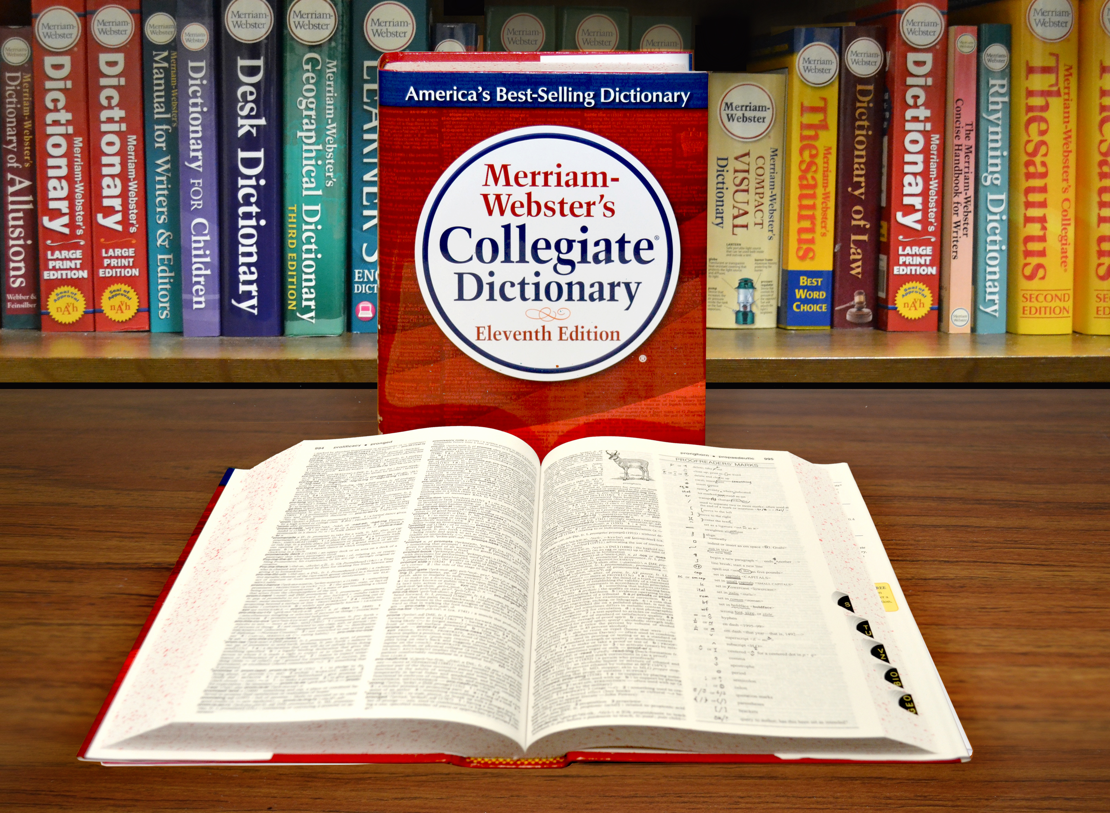 Merriam-Webster szótárak egy polcon, előttük a Collegiate Dictionary borítója, és egy közepén kinyitva lefektetett példány